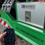 A hand holding a remote control in front of the GreenLoader control box.