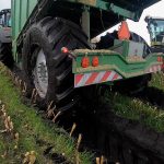 Low rear view of a GreenLoader in deep mud, with a forage harvester beside it.