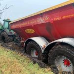 A wire rope runs from a tractor toward a red Bredal spreader sunk in mud.